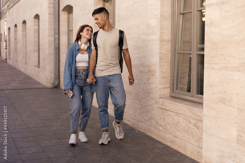 Full-length photo of young couple walking on street. Pretty dark-haired lady wearing denim outfit and white sneakers smiling and holding hand boyfriend in jeans and t-shirt