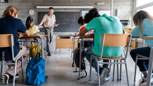 Mature caucasian man teacher hands out exams to multiracial high school students. Students ready to take exam. Horizontal photo banner