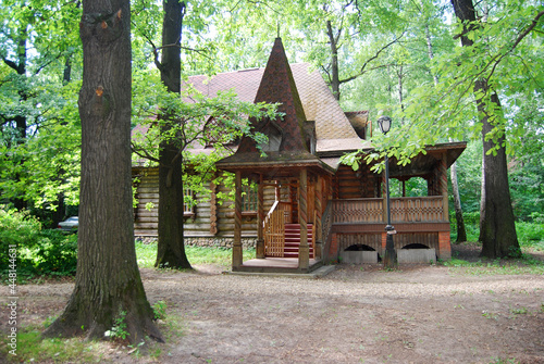 A wooden log hut with a patterned porch among green trees in the forest. An old log house among the trees. A porch with a triangular roof. Architecture