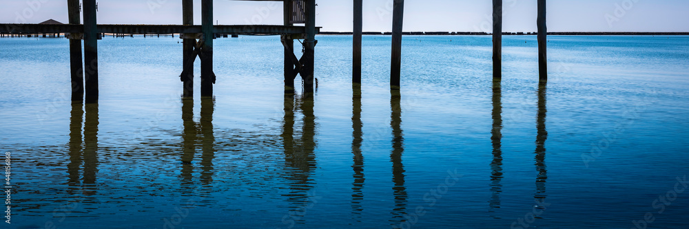 Under the boardwalk over blue seawater. Crossing geometry of vertical pilings and stretching jetty.