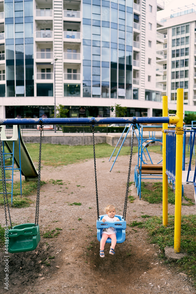 Little girl sits on a swing on a playground in the courtyard of an apartment building