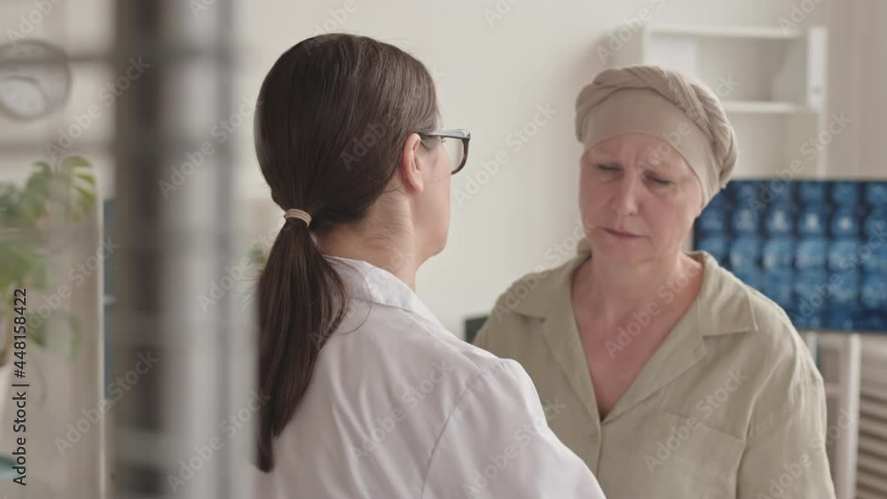 Panning medium close-up with slowmo of supporting female doctor hugging sad mid-adult woman suffering cancer standing together at doctor office Panning medium close-up with slowmo of supporting female doctor hugging sad mid-adult woman suffering cancer standing together at doctor office