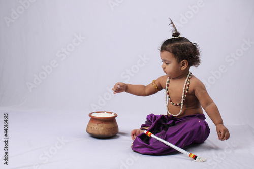 adorable Indian baby in krishna kanha or kanhaiya dress posing with his flute and dahi handi (pot with curd) on white background. sitting pose photo