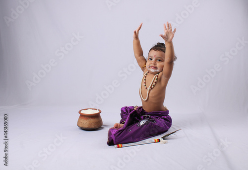 adorable Indian baby in krishna kanha or kanhaiya dress posing with his flute and dahi handi (pot with curd) on white background. sitting pose photo