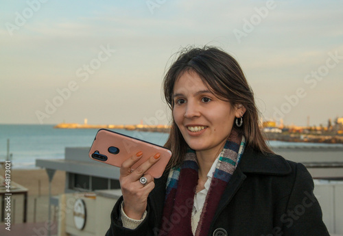 Young smiling woman talking on cell phone outdoors on the sea coast in  an autumn sunset © Ruben