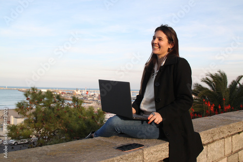 Autumn. Young sheltered woman on the coast checking emails on a laptop on a cool sunny day. she is happy and smiling © Ruben