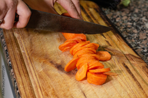hands cutting an carrot into slices on a wooden board with other vegetables. A fresh meal is being cooked © Ruben