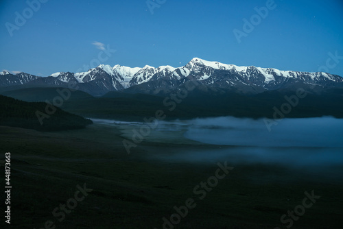 Atmospheric landscape with dense fog on lake and great snowy mountain ridge under night sky. Alpine scenery with thick fog on mountain lake and big mountain range in night. Snow pinnacle in dusk.