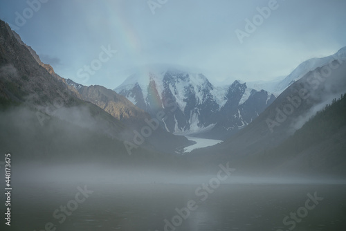 Atmospheric alpine landscape with mountain lake in fog and snowy mountains with rainbow in rainy weather. Gloomy scenery with mountain lake with rainy circles in fog on water and low clouds in valley. © Daniil