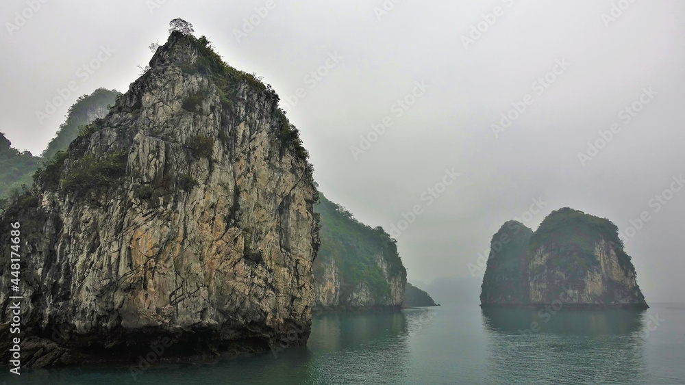Islands with bizarre outlines and steep slopes are hidden in the fog. Reflection on the surface of the calm water of Halong Bay. Vietnam