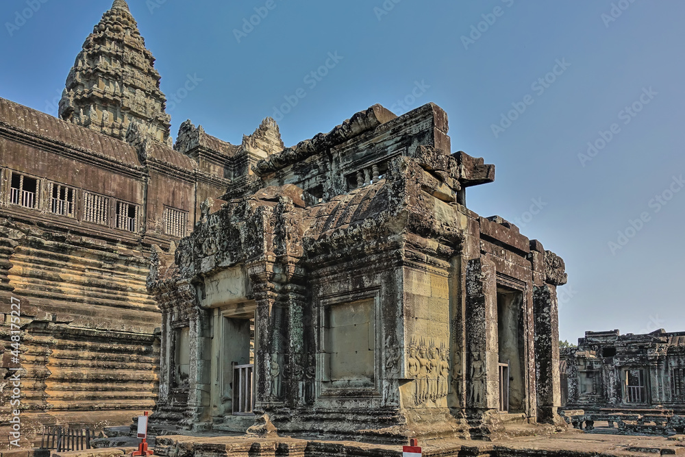The ruins of an ancient temple in the famous Angkor. Ornaments and bas-reliefs are visible on the weathered stone walls. A tall carved tower against a clear blue sky. Cambodia