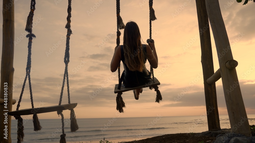 A dark silhouette of a girl on swing at seaside. A beautiful vacation leisure on a beach at sunset near ocean shore. Tropical romance and enjoyable time swinging alone.Anonymous female flying on swing