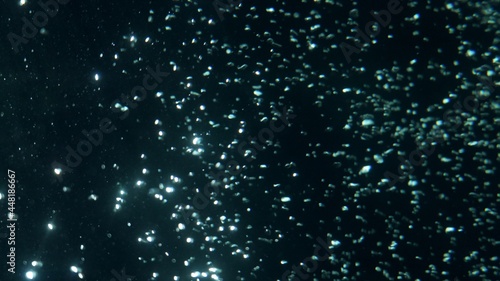 A beautiful texture of oxygen bubbles glowing in the dark water. Cosmic landscape with glittering particles and slow motion movement. Blue green spheres glistening in light on black space backdrop.