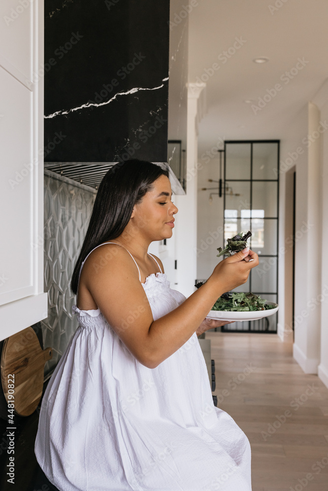 Pregnant woman sitting on counter in kitchen eating healthy green salad Pregnant woman sitting on counter in kitchen eating healthy green salad