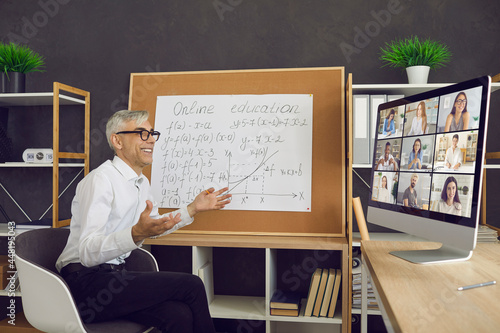 Happy mature man teaching math via video call on desktop computer. Smiling male teacher sitting in chair in front of classroom board having virtual online class with group of distant college students