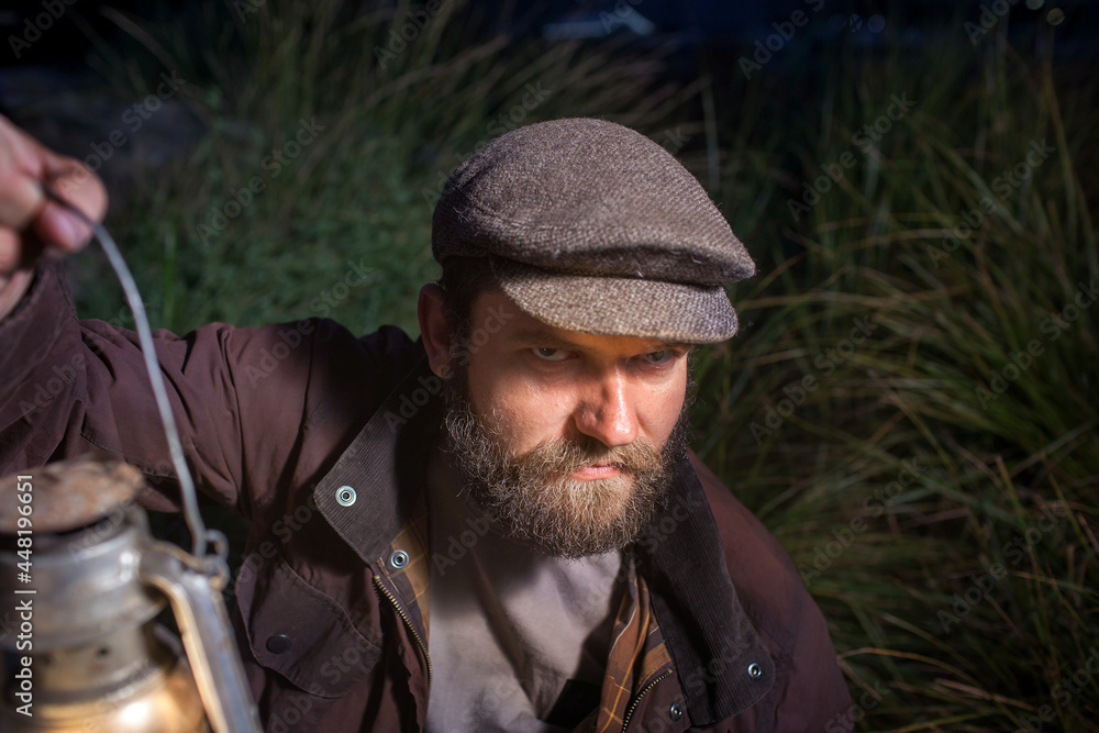 a man  with an evil confident face with a beard and in vintage clothes holds a kerosene lamp in his hands. It's dark around