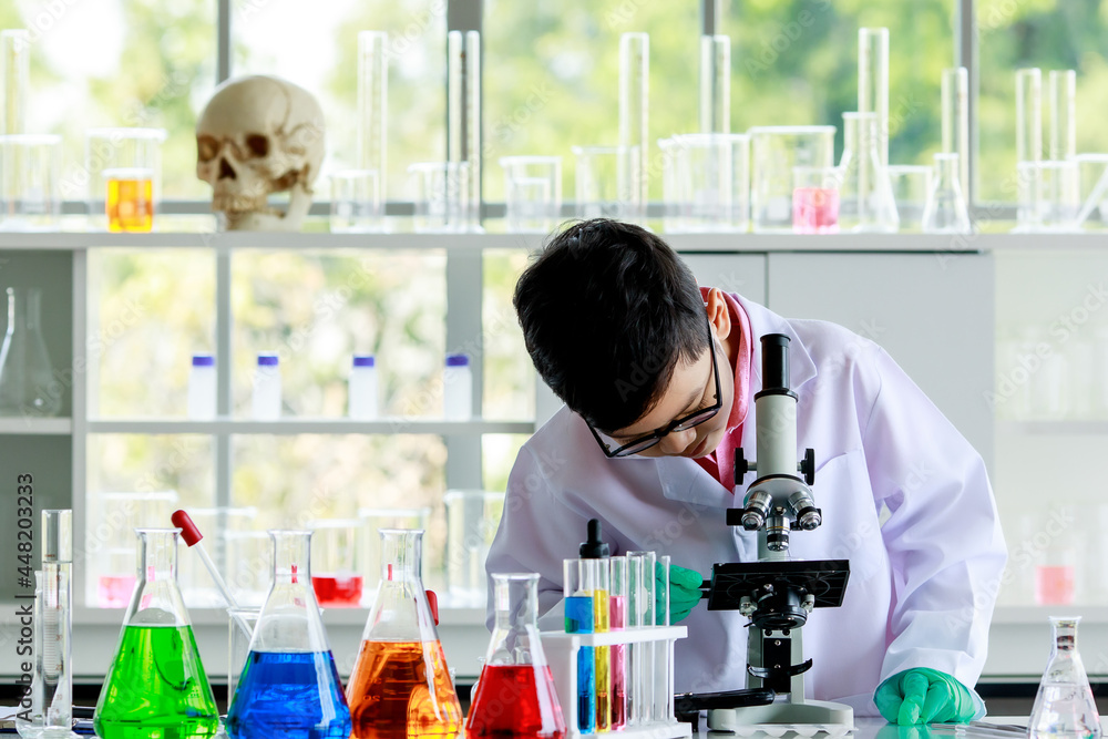 Asian schoolboy in glasses and lab coat examining chemical substance through microscope while conducting scientific experiment in school laboratory Asian schoolboy in glasses and lab coat examining chemical substance through microscope while conducting scientific experiment in school laboratory