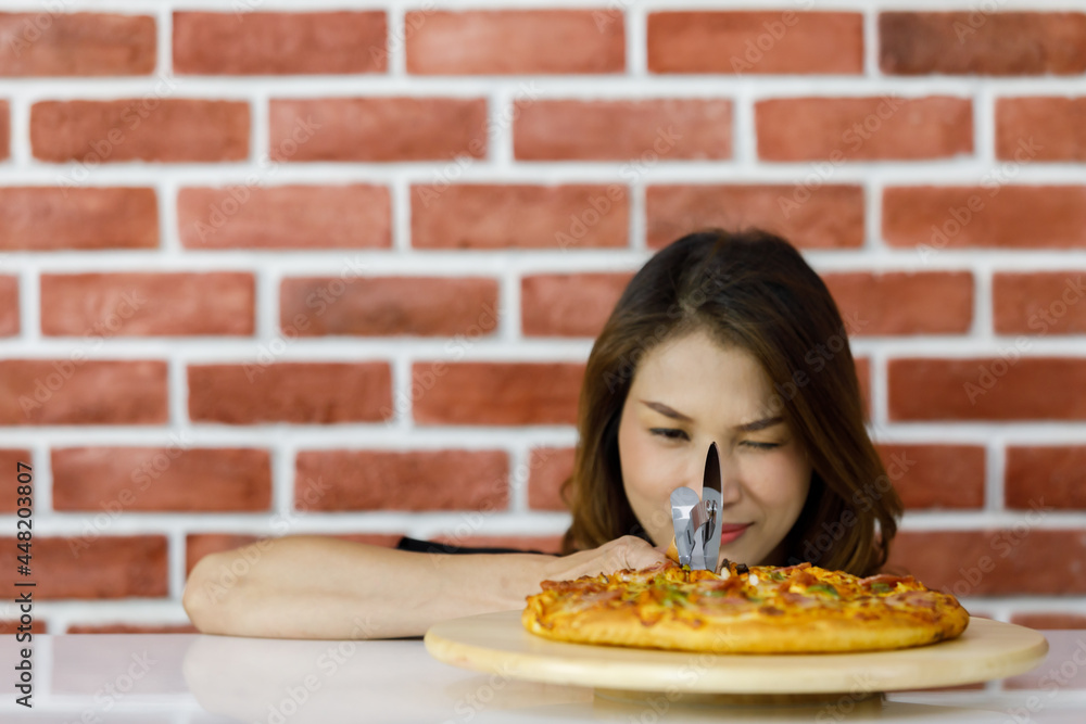 Beautiful Asian woman lower down near brick wall and smilingly looking very close at attractive tasty and healthy fresh pizza as her loved Italian food on round wooden plate on white table