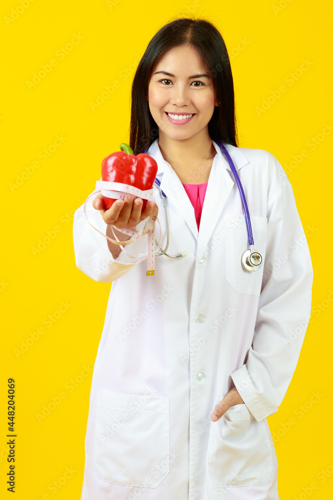 Asian young female doctor wearing white gown uniform with stethoscope, smiling, holding, presenting vitamin capsule pills with isolated yellow background and copy space. Nutrition, Healthy Concept. Asian young female doctor wearing white gown uniform with stethoscope, smiling, holding, presenting vitamin capsule pills with isolated yellow background and copy space. Nutrition, Healthy Concept.