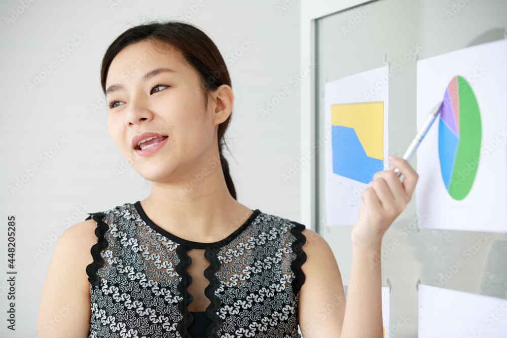 Asian young pretty successful friendly female businesswoman wears black gray white sleeveless dress stand looking and using pen pointing at chart and graph paper document presentation on glass wall Asian young pretty successful friendly female businesswoman wears black gray white sleeveless dress stand looking and using pen pointing at chart and graph paper document presentation on glass wall