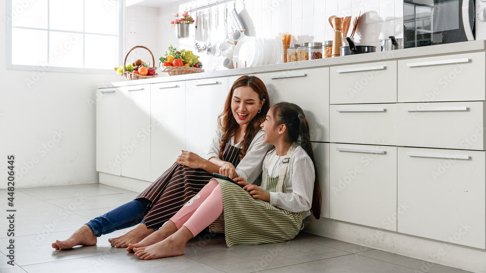Asian mother and daughter happily sit and study how to make cakes from their laptops and smile in the kitchen at home.. Asian mother and daughter happily sit and study how to make cakes from their laptops and smile in the kitchen at home..