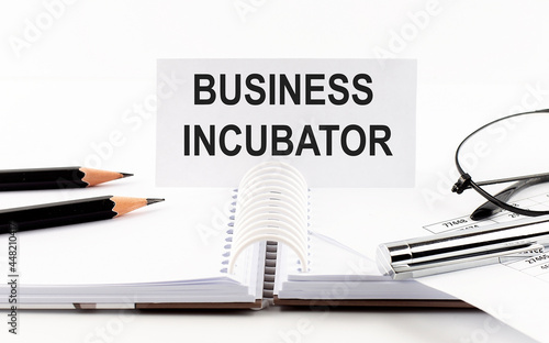 Text BUSINESS INCUBATOR on paper card,pen, pencils, glasses,financial documentation on the table - business concept