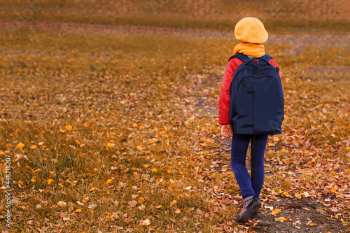 Autumn pittle girl in a yellow beret on a background of autumn glade with fallen leaves. A child with a backpack goes to school © Maryana