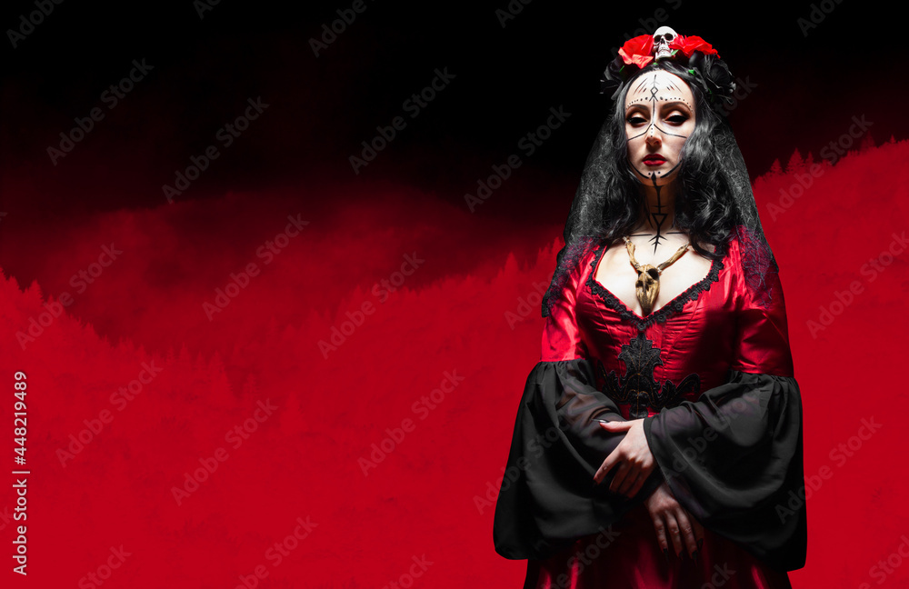 Mysterious woman, witch with black eyes and runic makeup in red gothic dress, crown with skull and roses and wooden animal skull amulet standing in crimson colored stylized forest. Halloween concept.