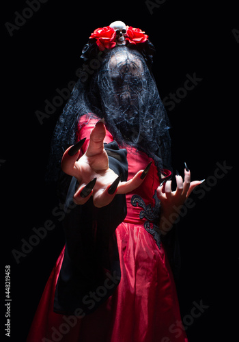 Photo Witch in gothic dress, black bridal veil and crown with skull and roses conjuring with her hands from the dark