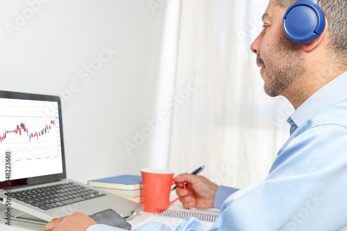 Hispanic man reviewing his investments at home on his laptop drinking coffee and listening to podcats photo