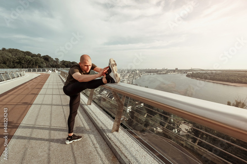 Fototapeta Naklejka Na Ścianę i Meble -  Young European man stretches his legs on footbridge in the city. Healthy lifestyle concept. High quality photo