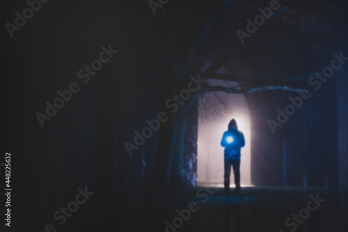 A spooky hooded man holding a glowing light  standing underneath a street light  On a foggy winters night. With a blurred  textured edit.