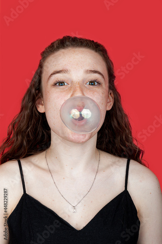 Redhead teenage girl makes a bubble gum bomb while looking at you with red background. Concept portraits and bubble gum