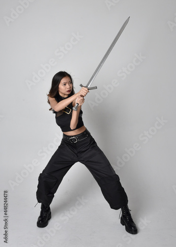  Full length portrait of pretty young asian girl wearing black tank top, utilitarian pants and leather boots. Standing pose holding a sword, isolated against a studio background.