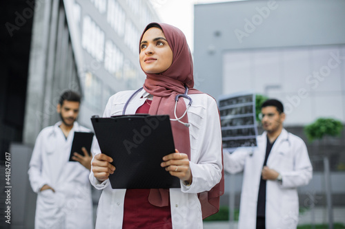 Canvas Print Pretty smiling Arab female doctor in hijab poses outdoors in front of modern clinic and looks away