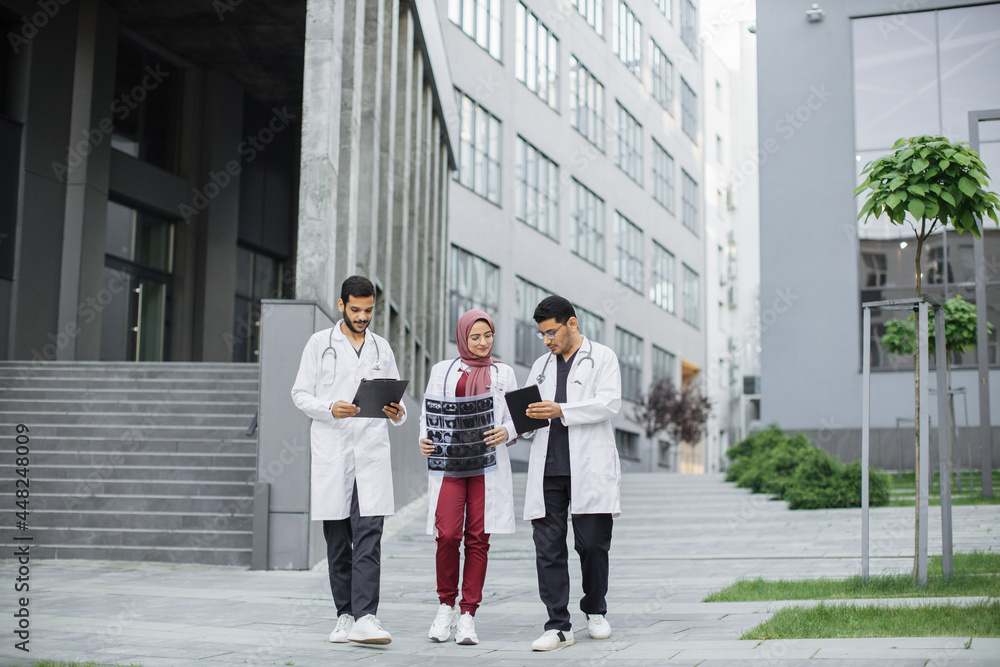 Obraz premium Three Arabian Muslim doctors, in medical suits and coats, discussing patient's x-ray tomography scan, walking outside on the background of modern hospital with stairs