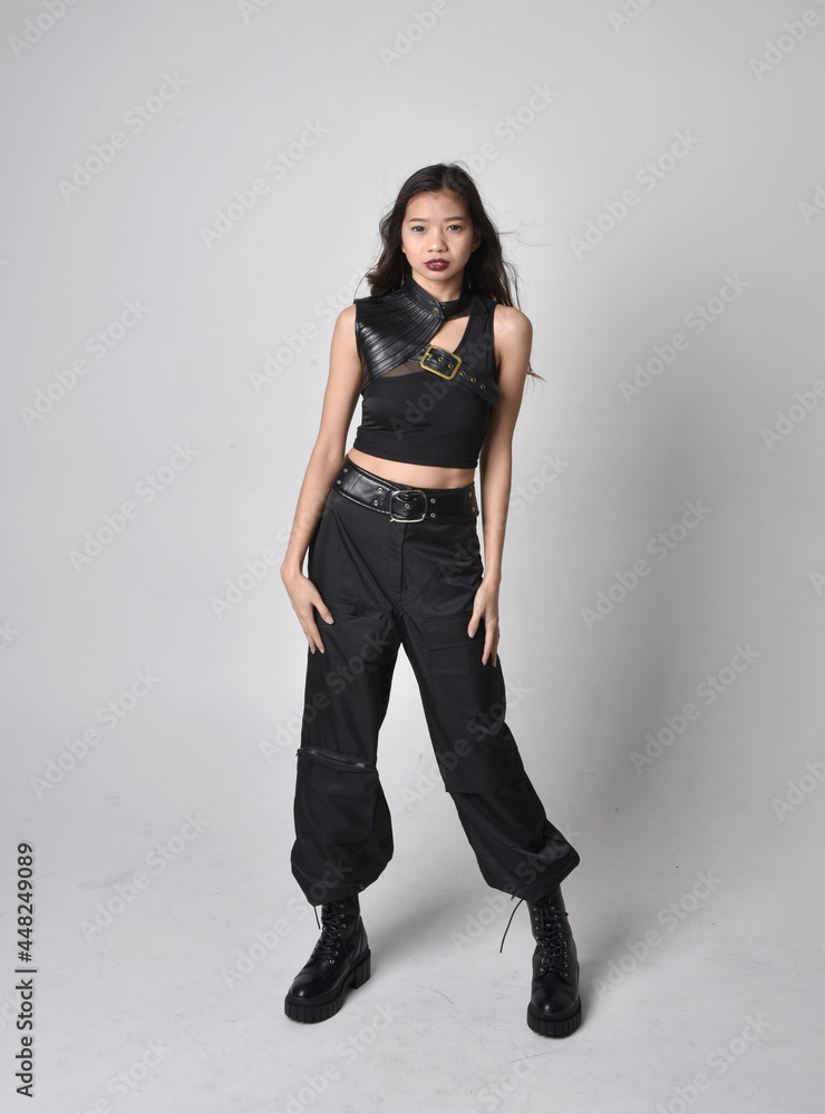 Full length portrait of pretty young asian girl wearing black tank top, utilitarian  pants and leather boots. Standing pose with gestural hands, isolated against a  studio background.