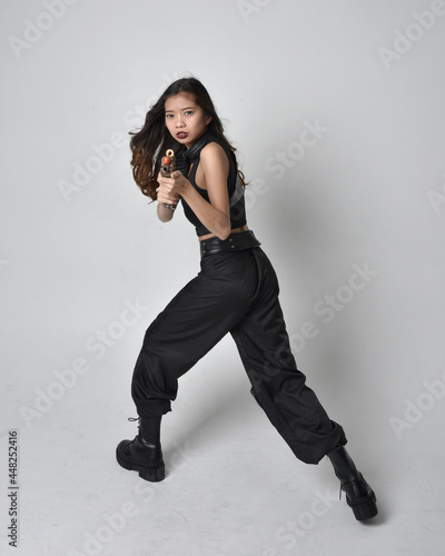 Full length portrait of pretty young asian girl wearing black tank top, utilitarian pants and leather boots. Standing pose holding a gun, isolated against a studio background.