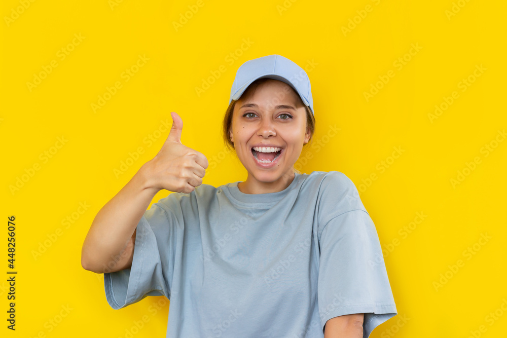 A young caucasian beautiful cheerful smiling blonde woman in a blue cap and oversize t-shirt showing thumb up like gesture with her hand isolated on a bright color yellow background A young caucasian beautiful cheerful smiling blonde woman in a blue cap and oversize t-shirt showing thumb up like gesture with her hand isolated on a bright color yellow background