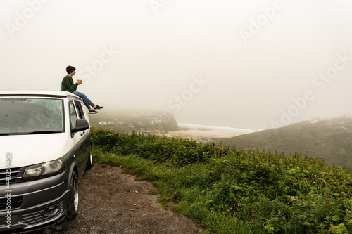 Attractive young female sitting on the roof of a campervan while enjoying the views from the top of a cliff
