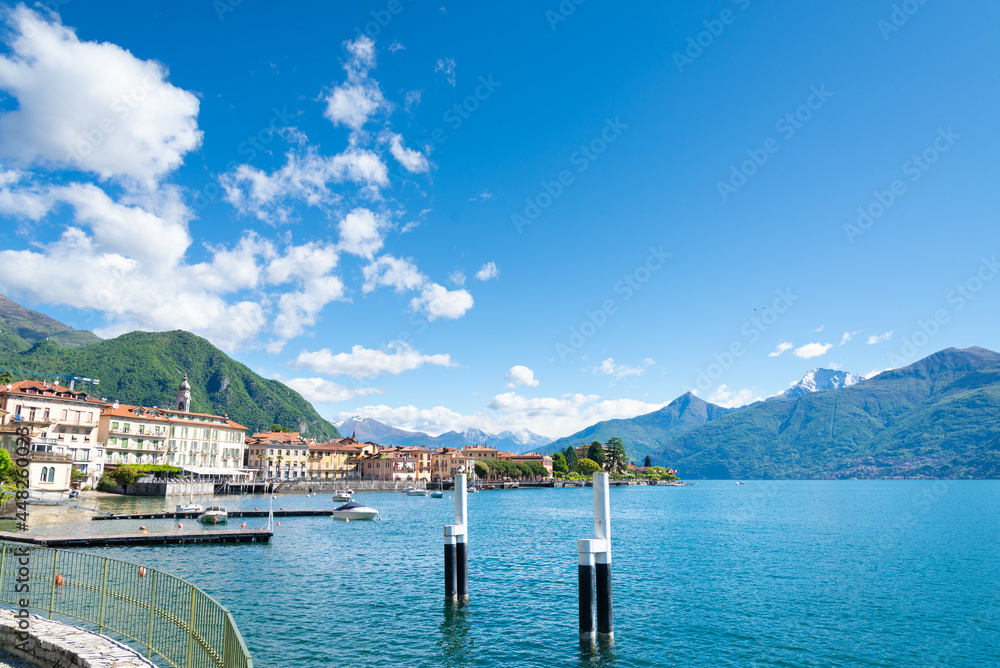 The small harbour of the village of Menaggio, Lake Como, Italy, with a view of the coast. Blue turquoise waters on the right. Italian alps, blue sky and clouds on the background.