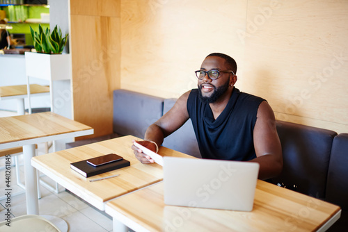 Happy dark skinned male blogger with touch pad and laptop technology spending weekend leisure in cafe interior enjoying time for web networking, smiling African American IT professional in glasses