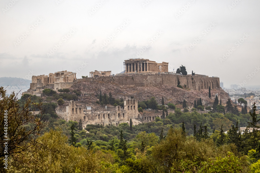 Acropolis hill (Parthenon, Propylaea, Temples, Odeon of Herodes Atticus) in summer greenery. Athens ancient historical landmark in city center from Filopappou Hill on cloudy day
