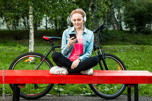A girl with a bicycle sits on a park bench and listens to music with headphones.