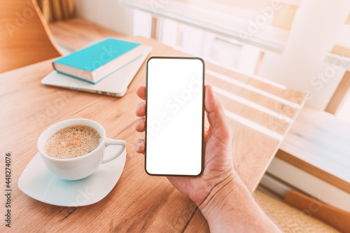 Blank mobile smart phone screen mock up. Man holding smartphone over desk with coffee cup at home.