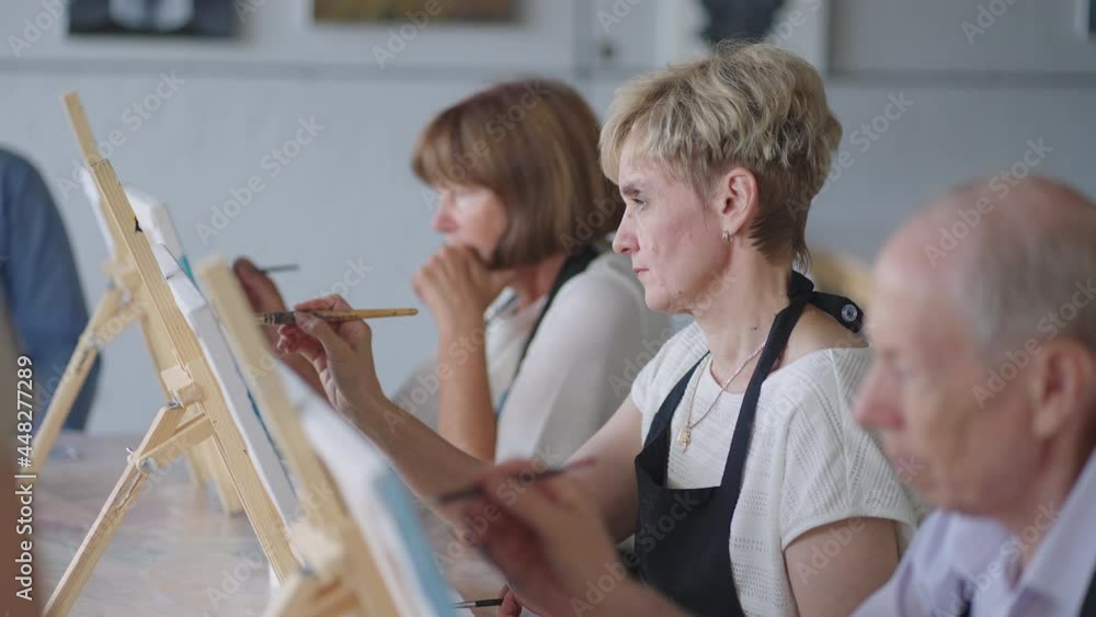 Side view of a happy senior people smiling while drawing as a recreational activity or therapy in paint class together with the group of retired women and men Side view of a happy senior people smiling while drawing as a recreational activity or therapy in paint class together with the group of retired women and men