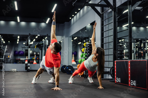 The fitness man and energetic woman are in a plank position with their arms raised and doing full-body in a modern gym. Body stability, Motivation, better together