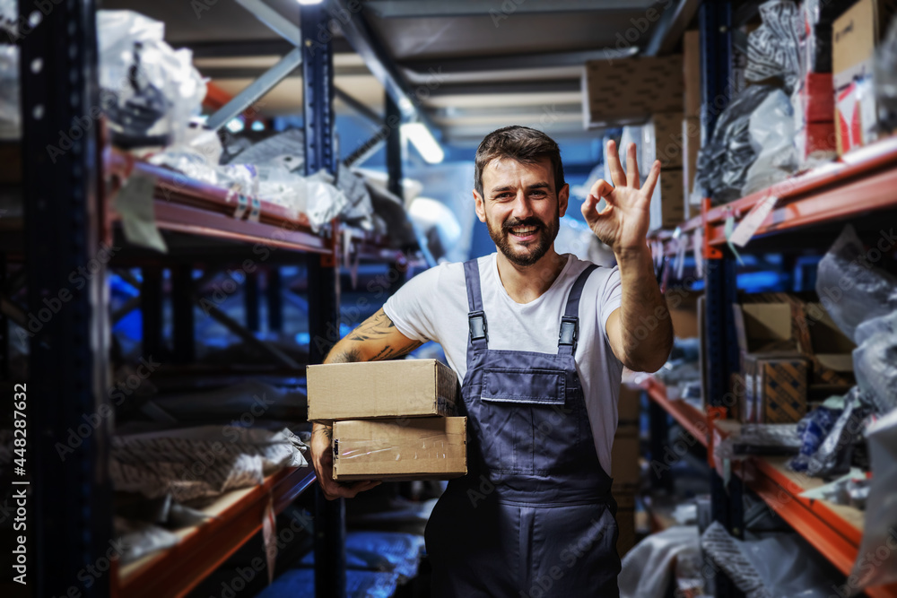 Young smiling happy bearded tattooed hardworking employee holding boxes and standing in storage of import and export firm and showing okay gesture.