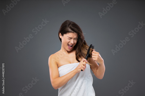 Painful combing of tangled hair with hairbrush. Woman wrapped in a white towel around her body stands in front of a gray wall and uses a hair comb after showering. Time for a haircut