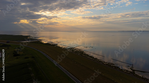 Luftaufnahme von einem Deich zwischen der Nordsee und einem ruhigem Gewässer im Sonnenuntergang mit einigen Wolken